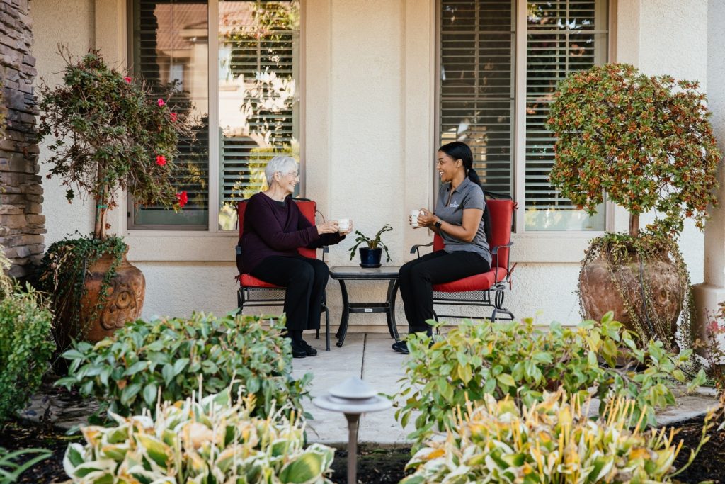 A caregiver and an older woman sit on a front porch surrounded by plants, enjoying coffee together on red patio chairs.