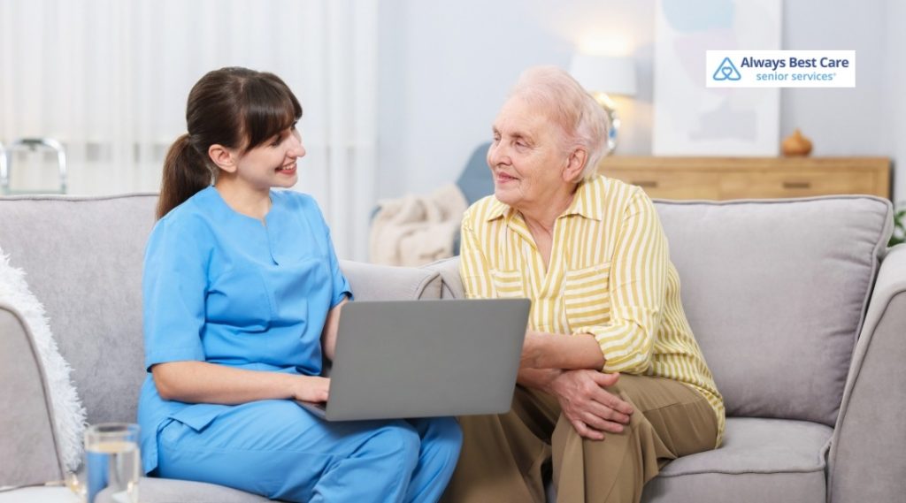 Smiling in-home caregiver sitting on a couch with an older adult, reviewing information together on a laptop.