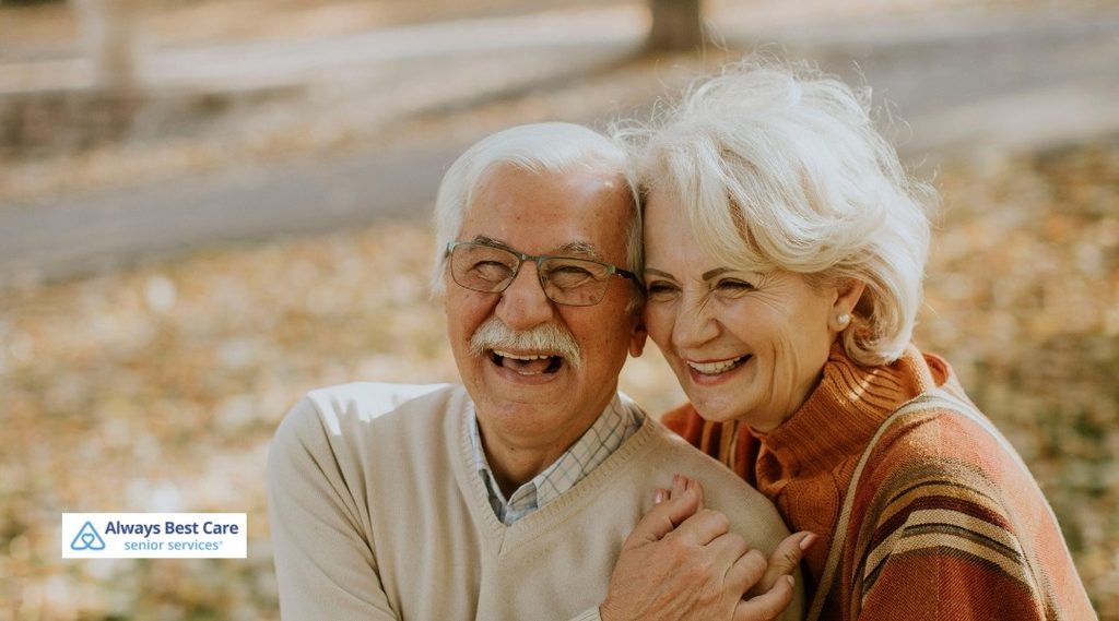 Senior couple smiling and embracing outdoors with autumn leaves in the background.
