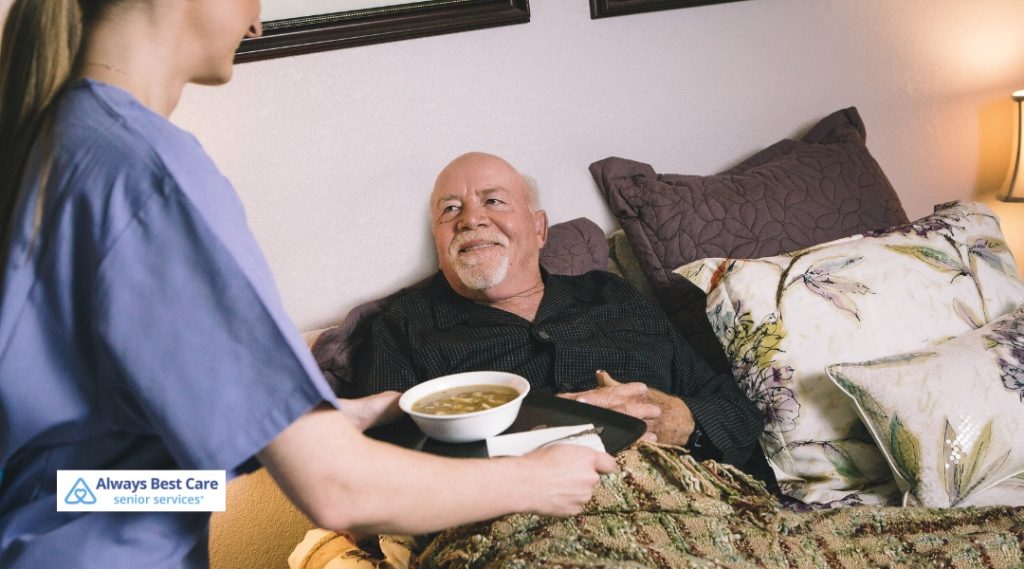 Caregiver bringing a bowl of soup to an elderly man lying in bed, both smiling at each other, with soft lighting in a cozy room.