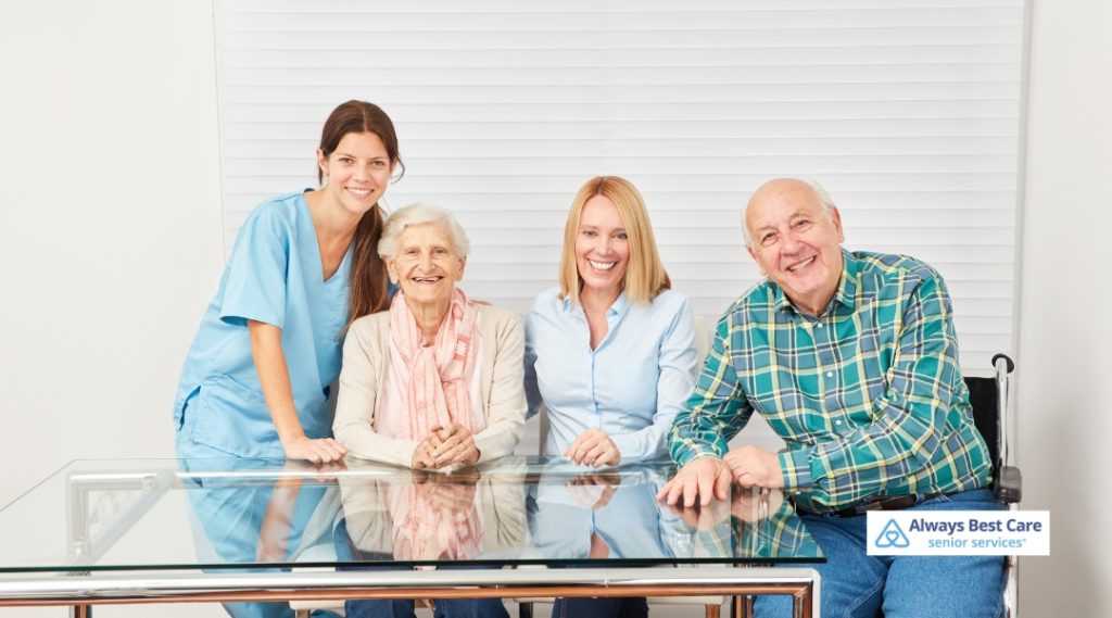 A happy group portrait featuring a female caregiver, two senior adults, and a family member sitting together at a glass table, all smiling. The Always Best Care logo is present in the image.