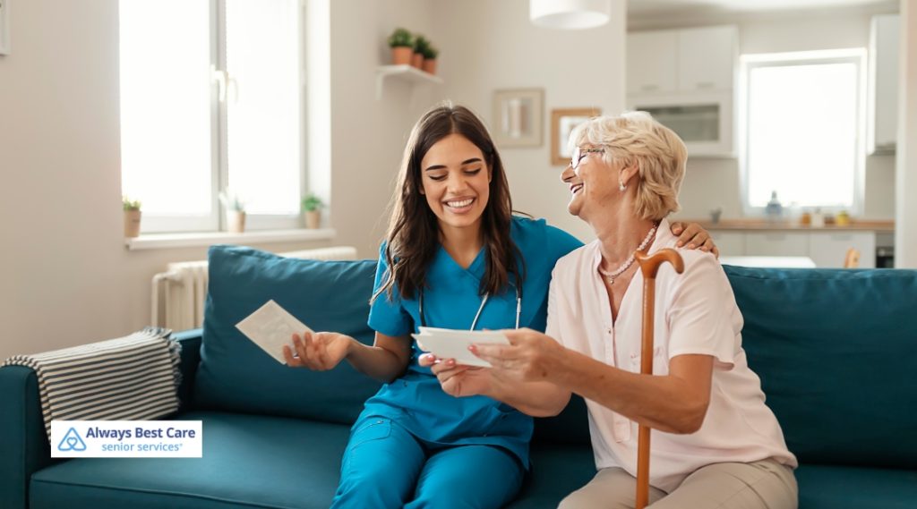 A young caregiver in blue scrubs and an older woman with a cane sit on a blue couch, laughing while looking through photographs. The Always Best Care logo is visible in the bottom left corner.