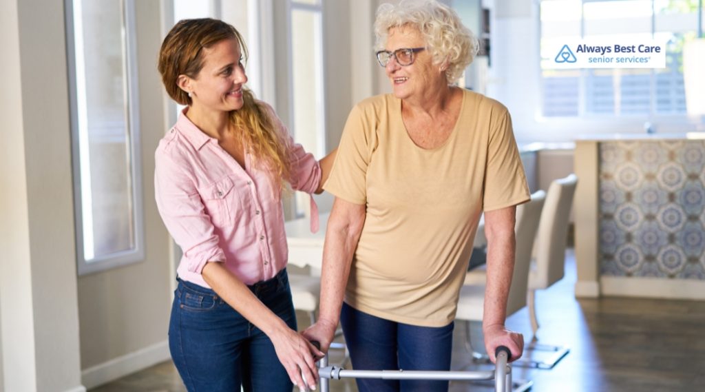 A caregiver is assisting an elderly woman with a walker in a well-lit room. The caregiver is gently guiding the woman, who is smiling as they walk together.