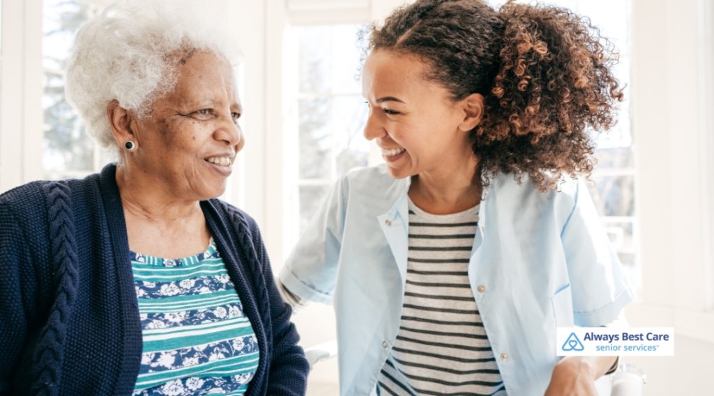 A caregiver is assisting an elderly woman in the home, both sharing a cheerful moment, with the elderly woman laughing and the caregiver supporting her with a warm and friendly expression.