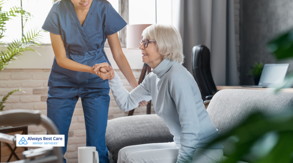 A caregiver assisting an elderly woman in standing up from a couch in a well-lit living room. The caregiver is offering her hand to help the senior, ensuring her comfort and safety.