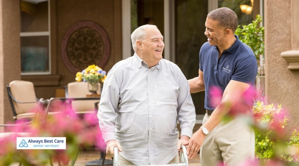A caregiver walks alongside a senior man, offering support as he uses a walker in a vibrant outdoor setting. They share a smile, showcasing the compassionate companionship Always Best Care provides.