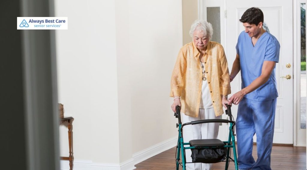 A caregiver assisting an elderly woman with a walker as they walk together through a bright and welcoming home environment, ensuring the senior’s safety and comfort.