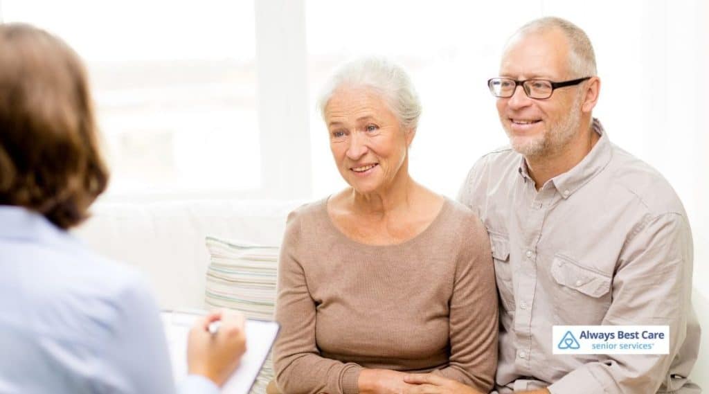 A senior couple sits together smiling while a caregiver takes notes, reflecting the compassionate care Always Best Care offers to ensure seniors' well-being.