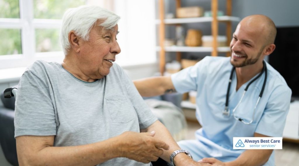 Caregiver sitting beside an older man at home, sharing conversation and emotional support
