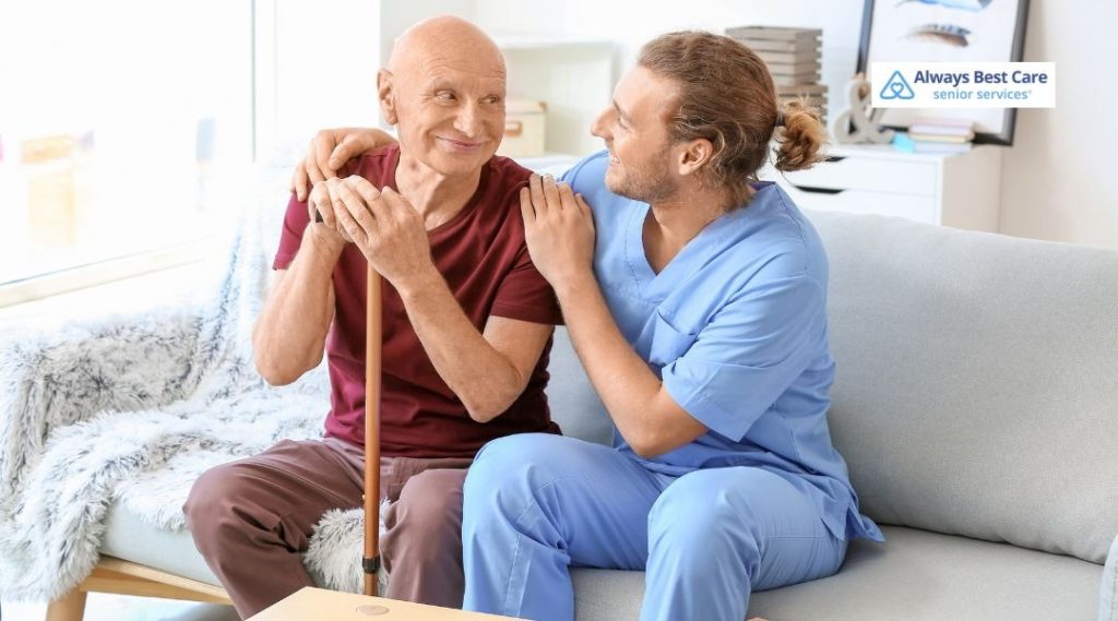 Caregiver in blue scrubs sitting close to an older adult with a cane on a sofa, resting a hand on their shoulder in a supportive gesture.