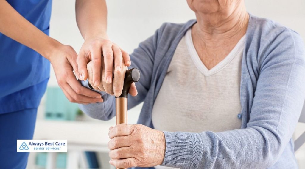 Caregiver in blue scrubs placing a supportive hand over an older adult’s hands on a cane, symbolizing stability and mobility assistance.
