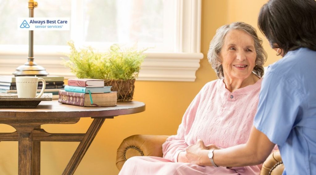 In-home caregiver in blue scrubs sitting beside an older adult in a cozy living room, holding their hands and offering comfort and conversation.
