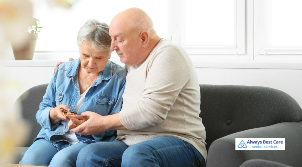 Older woman checking her blood sugar with support from her husband while sitting on a sofa.
