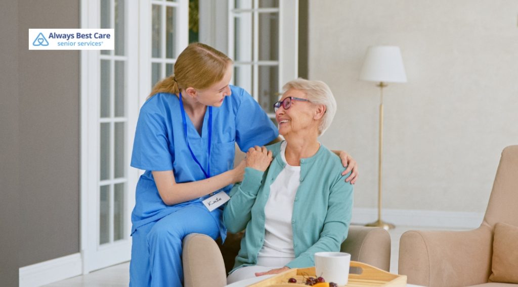 A young female caregiver in blue scrubs gently holds hands and smiles warmly at an elderly woman wearing glasses and a teal cardigan. They are seated indoors in a bright, cozy room with glass doors and a lamp. The Always Best Care logo is in the top-left corner.