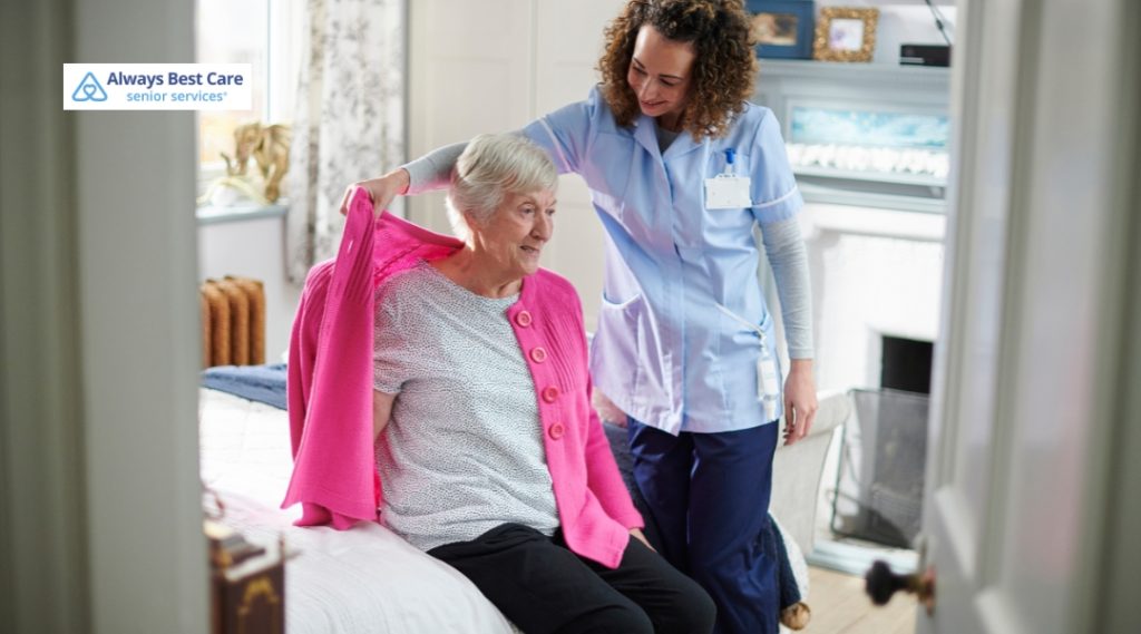 A female caregiver in light blue scrubs helps an elderly woman wearing a pink cardigan put on her coat. They are inside a warmly decorated bedroom with a fireplace. The Always Best Care logo is in the top-left corner.