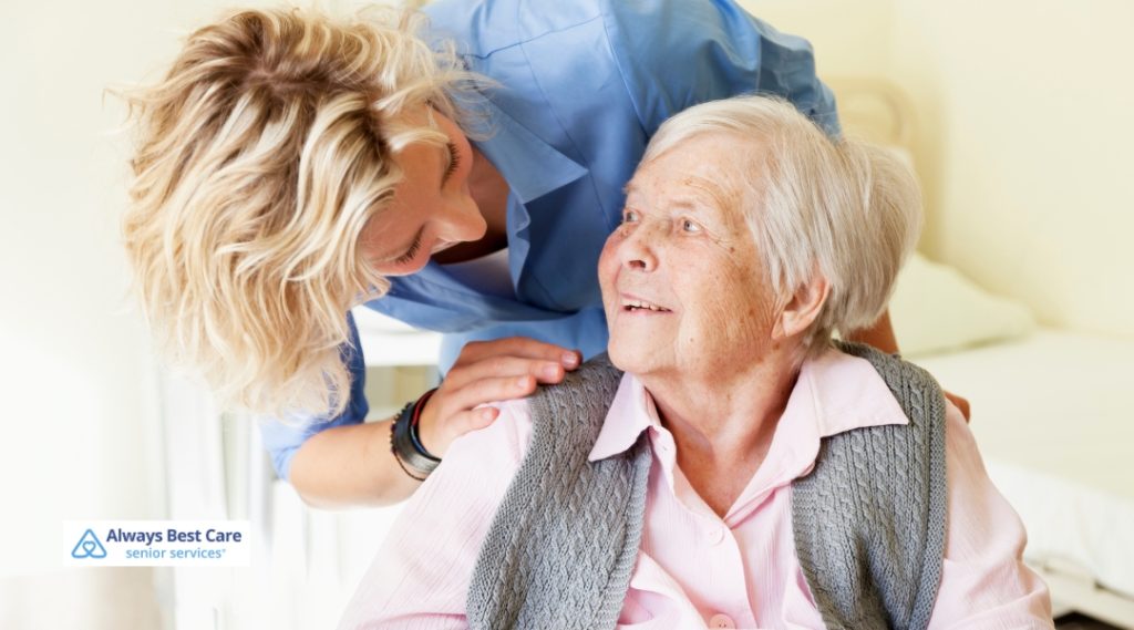 A female caregiver with blonde hair smiles affectionately at an elderly woman with short white hair and a gray sweater. They are indoors in a softly lit room. The Always Best Care logo is in the bottom-left corner.