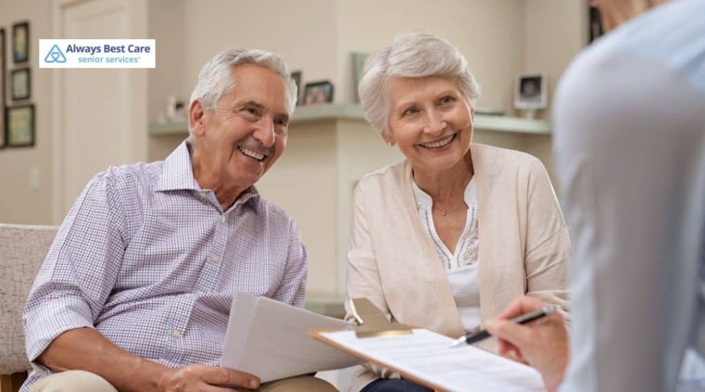 A senior couple sitting together with their caregiver, smiling while reviewing paperwork. The caregiver, a professional in a blue uniform, is engaging with the couple in a cozy, well-lit living room.