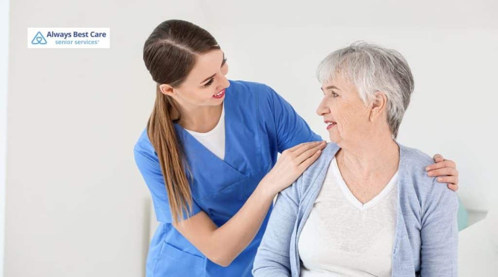 A senior woman and her caregiver sharing a warm, affectionate moment. The caregiver, a young woman in a blue medical uniform, is assisting the senior woman in a peaceful home environment.