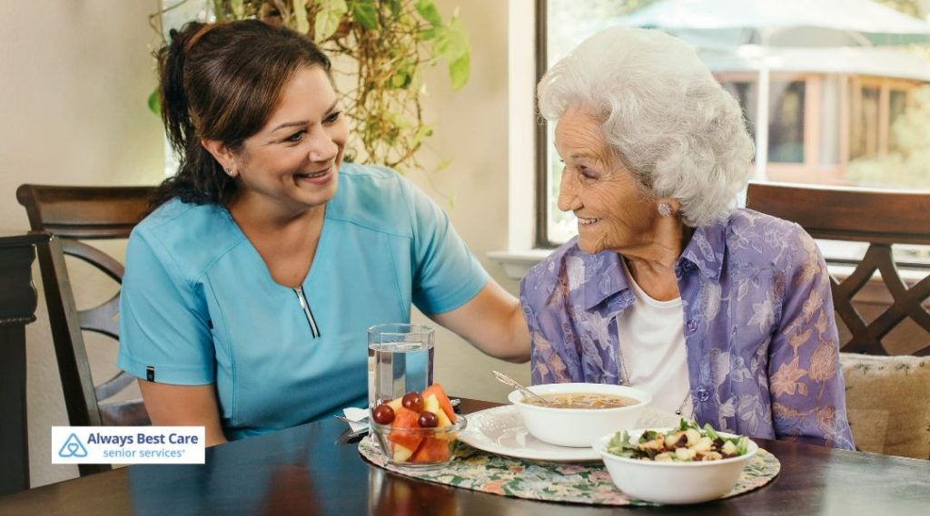 Caregiver serving soup, salad, fruit, and water to an older woman at the kitchen table.