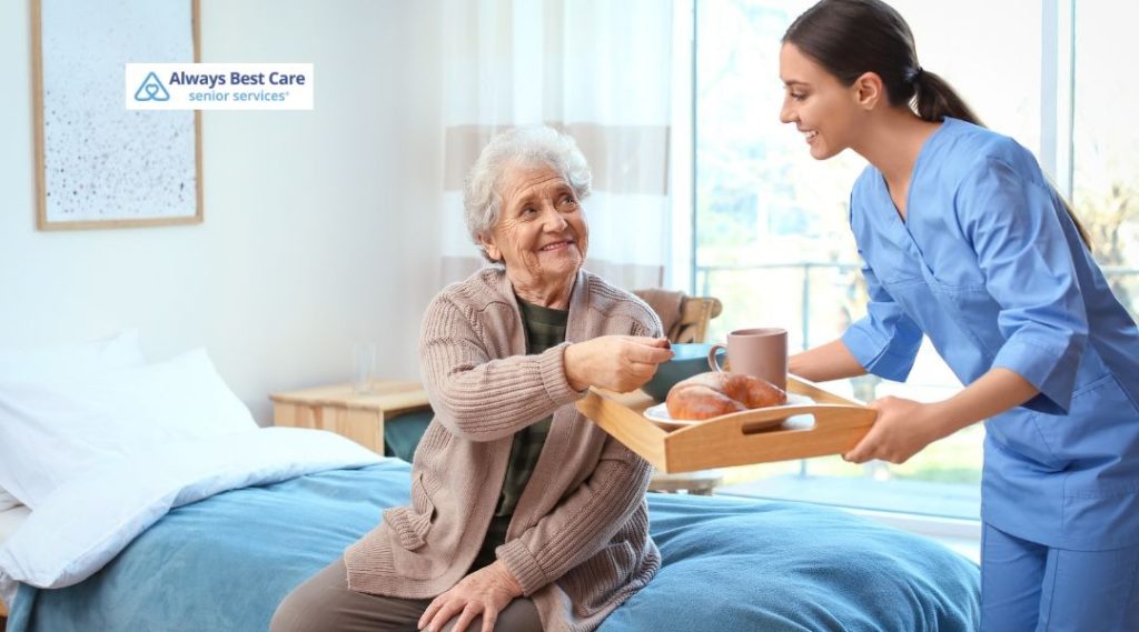 Caregiver serving breakfast on a tray to a senior woman sitting on the edge of her bed.