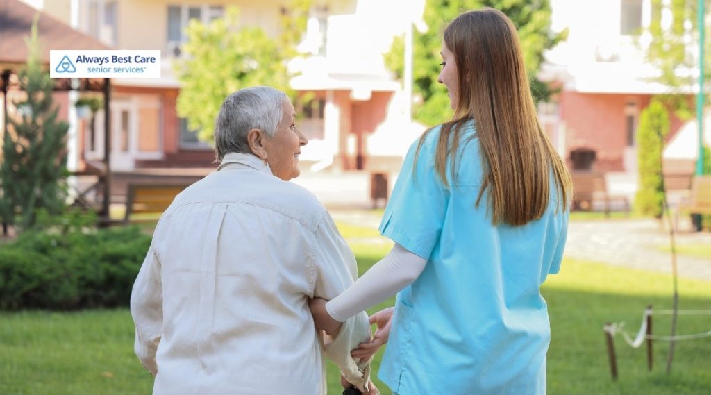 5. Young female caregiver in light blue scrubs walking hand-in-hand with an older adult through a green outdoor courtyard, both seen from behind, with the Always Best Care Senior Services logo in the corner.