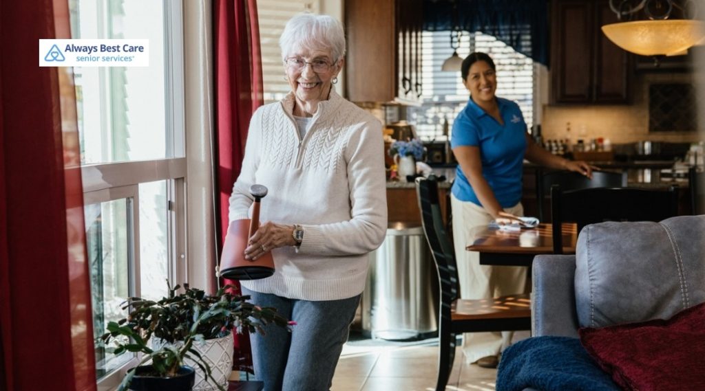 Smiling senior woman watering plants near a sunny window while a caregiver cleans in the background.