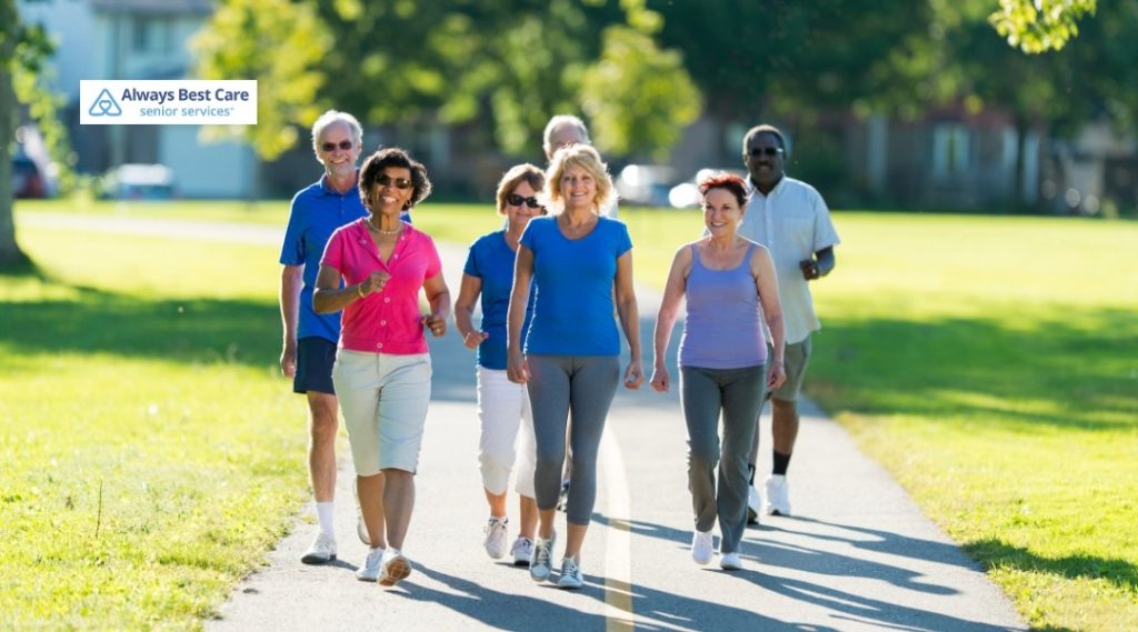 8. Group of active seniors walking together on a sunny park trail, wearing casual fitness clothing, with the Always Best Care Senior Services logo in the corner.