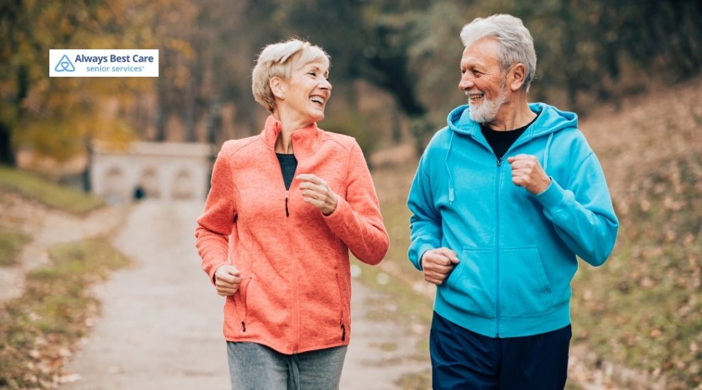 7. Older couple in bright athletic jackets power walking together on a tree-lined path in cool weather, with the Always Best Care Senior Services logo in the corner.