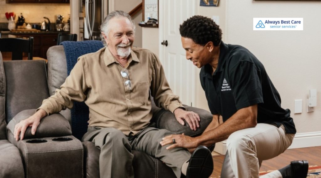 A caregiver kneels beside an older man seated in a recliner, gently supporting his leg as they work on mobility or transfer assistance in a cozy living room.