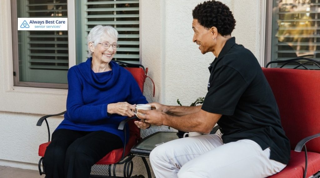 A caregiver hands a warm drink to an older woman seated on an outdoor patio, sharing a calm conversation in front of a home.
