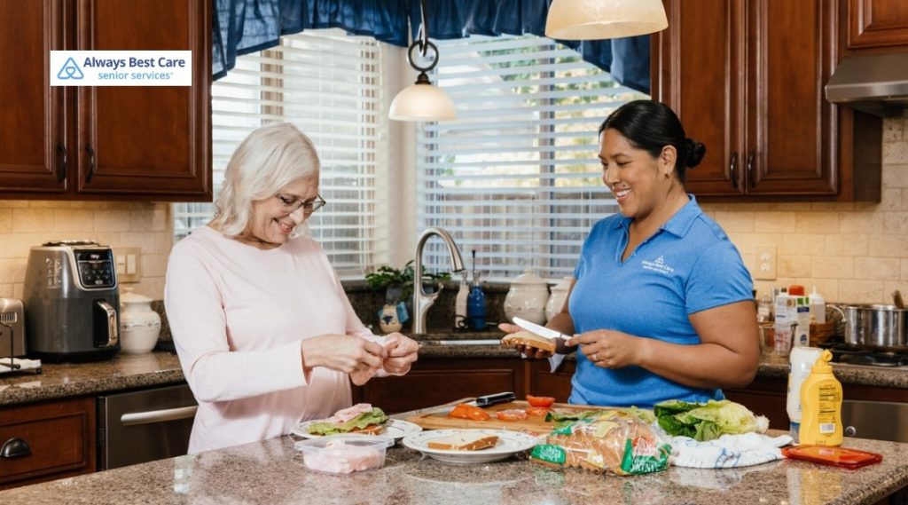 An older woman and a caregiver in an Always Best Care polo prepare sandwiches together in a bright kitchen, surrounded by fresh ingredients on the counter.