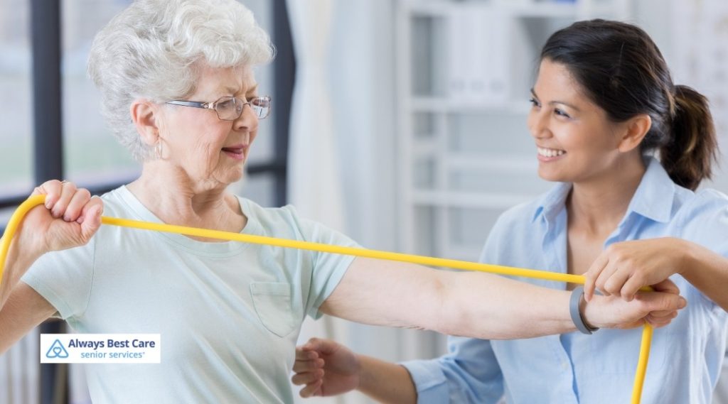 Senior woman stretching an exercise band with assistance from an Always Best Care caregiver during therapy.