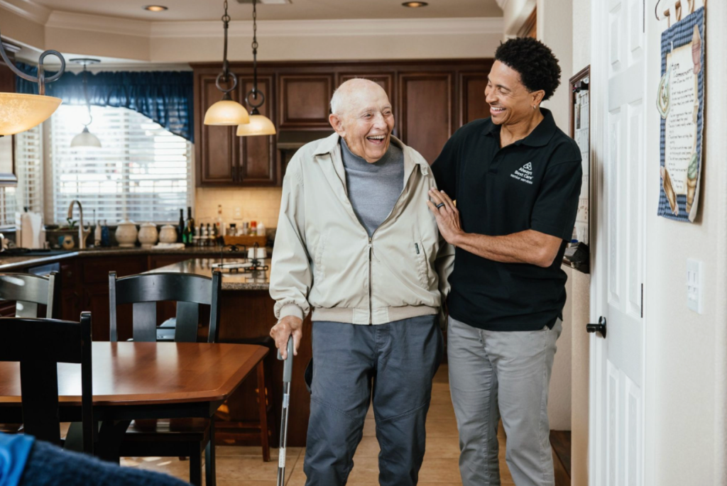 Caregiver supporting an older adult as he walks, with other seniors and staff visible in the background of a bright care setting