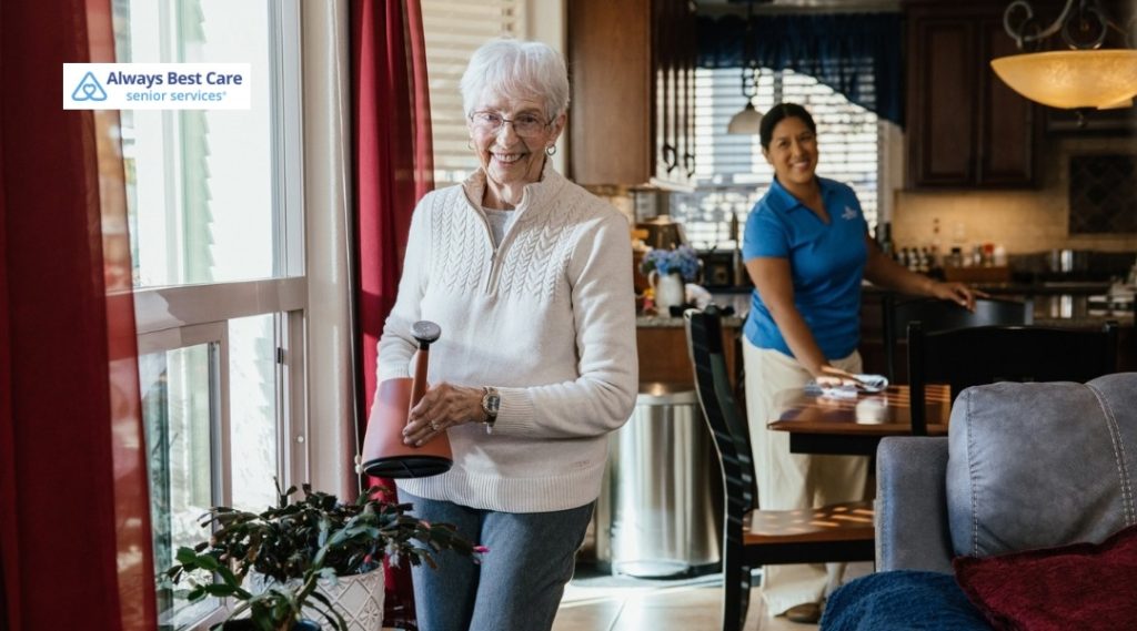 Older woman watering a houseplant by a bright window while a caregiver dusts and tidies the dining area behind her, with the Always Best Care Senior Services logo in the corner.
