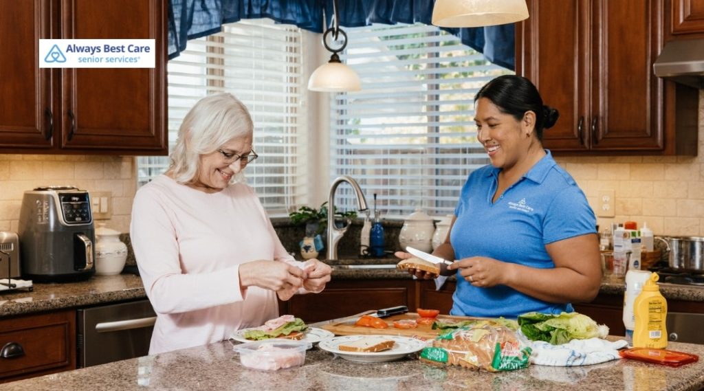 Older woman and caregiver standing together at a kitchen counter, preparing sandwiches with fresh ingredients laid out, with the Always Best Care Senior Services logo in the corner.