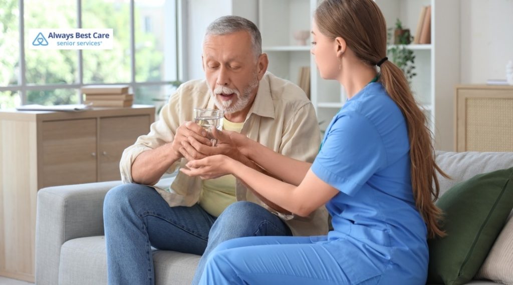 Female caregiver in scrubs helping an older man drink a glass of water while they sit together on a sofa.