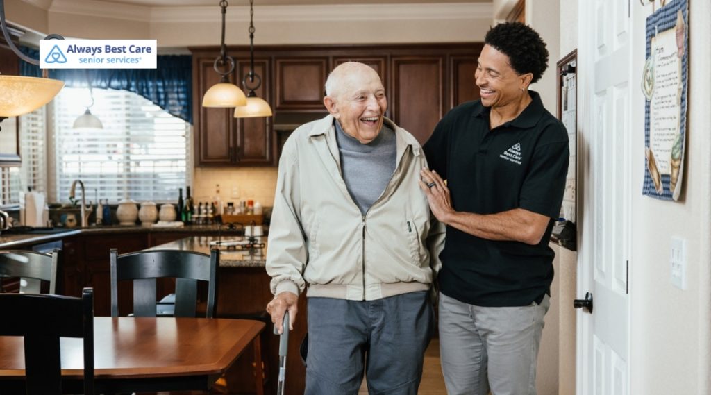 Caregiver supports a smiling older man as he walks through his home.