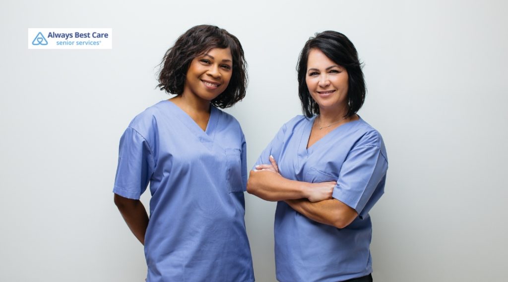 Two female caregivers in matching blue scrubs standing confidently against a white background, smiling and facing the camera.