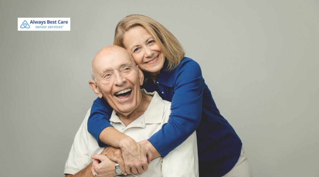 Smiling middle-aged woman hugging an elderly man from behind, both looking joyful and content in a close, affectionate pose.
