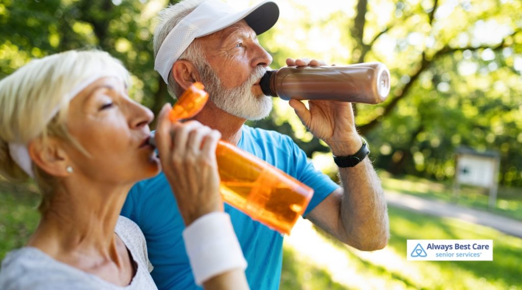 Elderly couple drinking water from sports bottles after exercising outdoors, surrounded by greenery.