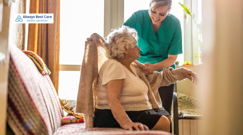 A caregiver helps an elderly woman put on a sweater, ensuring comfort and support. The elderly woman is seated in a cozy living space, with a bright and warm atmosphere.
