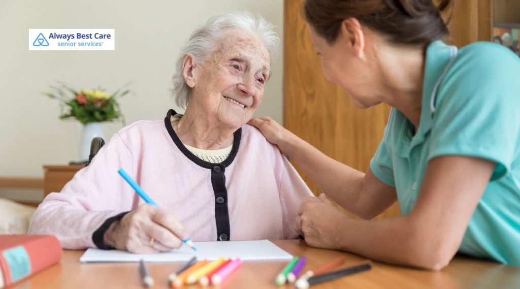 An elderly woman smiles brightly as she colors with crayons, while a caregiver gently places a hand on her shoulder, offering encouragement and companionship.