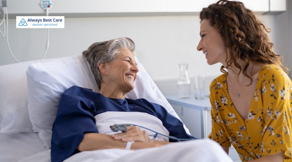 An elderly woman in a hospital bed smiles warmly at her visitor, who is holding her hand and offering companionship.