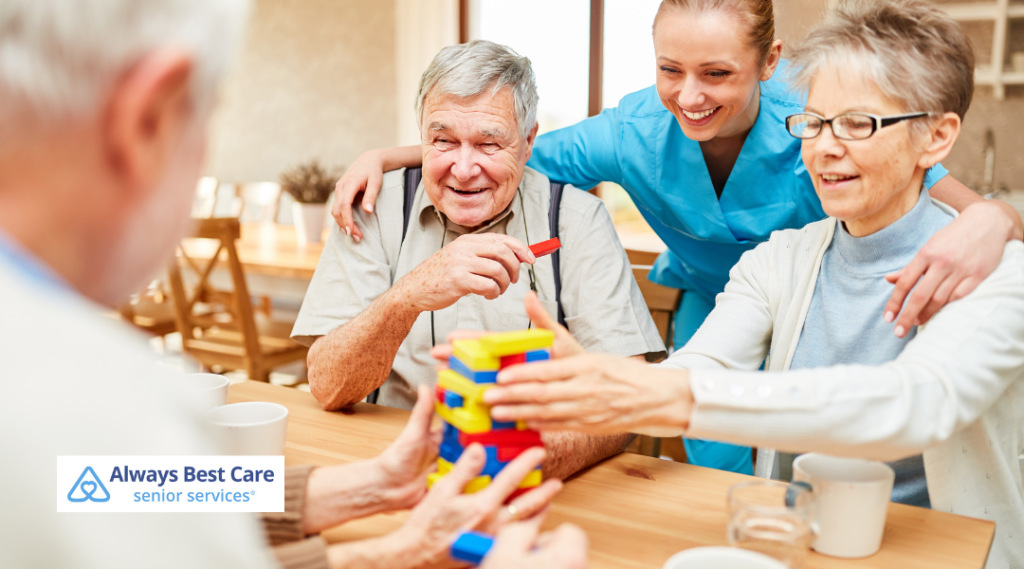 A caregiver and elderly individuals enjoying a fun game with building blocks, smiling and interacting in a comfortable indoor setting, with Always Best Care branding in the corner.