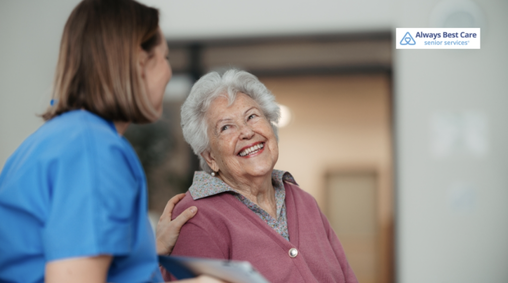 A senior woman laughing and looking up at her caregiver, who is holding a tablet. The caregiver is smiling back at her, and they are in a comfortable, indoor setting with soft lighting.