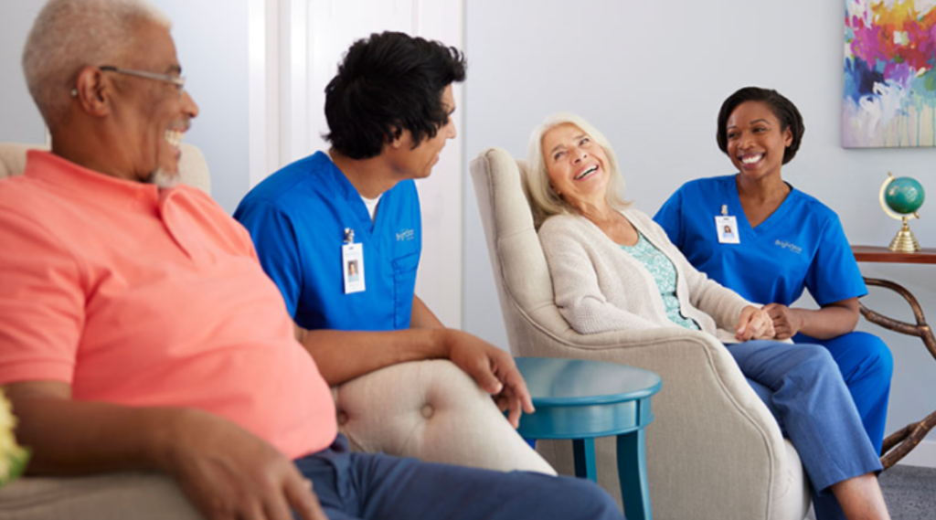 Two professional caregivers in blue uniforms sitting and talking with two older adults in a bright living room, creating a warm and supportive group setting.