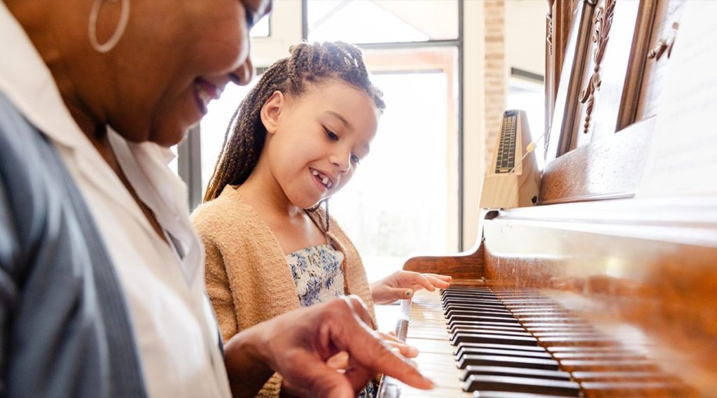 Senior lady t helping a child play the piano, their hands on the keys together, capturing a joyful intergenerational moment in a sunlit room.
