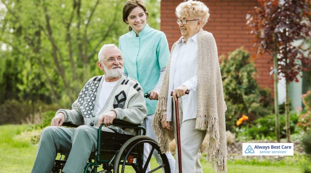 Caregiver standing behind an older man in a wheelchair beside a senior woman using a cane in a green yard.