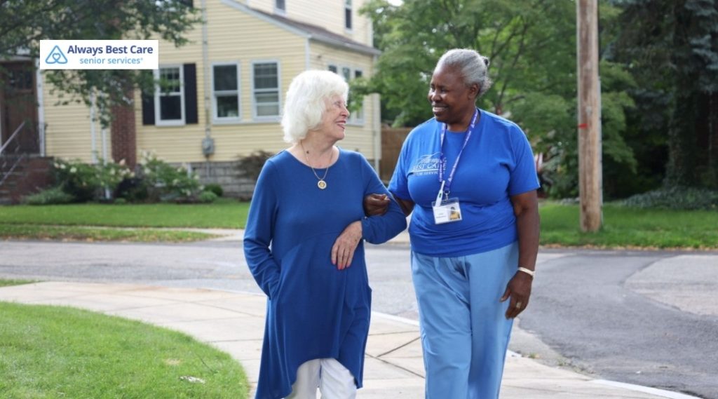 Always Best Care caregiver walking arm-in-arm with a senior woman along a quiet neighborhood sidewalk.