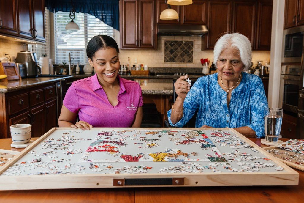 A smiling caregiver in an Always Best Care polo sits at a kitchen table helping an older woman assemble a large jigsaw puzzle.